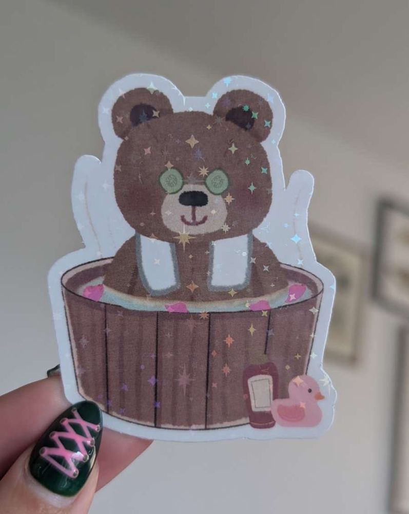 Holographic sticker of a bear in a bathtub, held between two fingers and catching the light